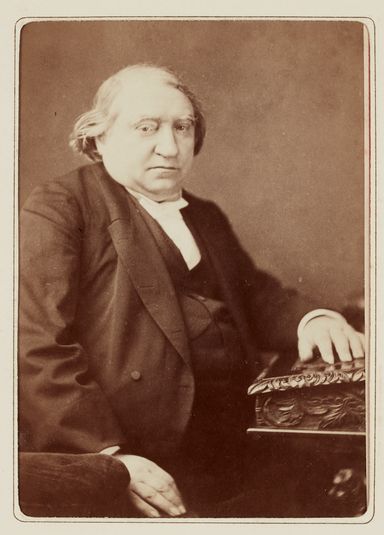 Ernest Renan, assis, main gauche posée sur un bureau, vers 1880