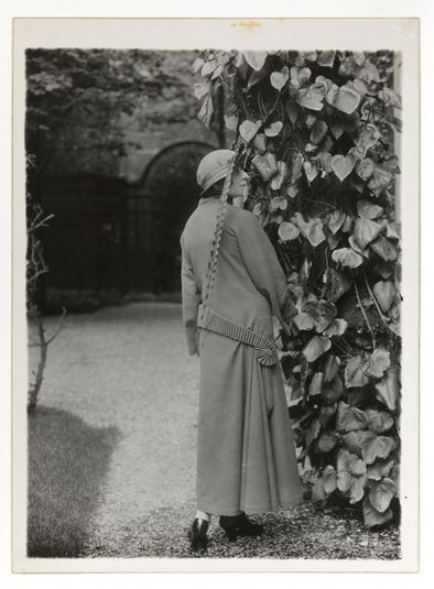 Modèle Chasse : modèle de dos avec manteau bordé de tissu tuyauté et drapé sur le côté et chapeau à longues plumes tombantes, maison Paul Poiret